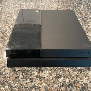 Puede incluir: Una consola PlayStation 4 negra, con un diseño rectangular. La consola tiene un acabado negro brillante en el lado izquierdo y mate en el derecho. El logotipo de PlayStation es visible, junto con la designación "PS4".