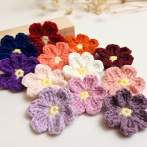 May include: A selection of handmade crochet flowers in shades of blue, purple, orange, pink, and white. Each flower features a yellow centre. The flowers are displayed on a white surface, indicating a craft or decorative purpose.