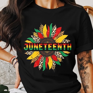 Puede incluir: Camiseta negra con un diseño de girasol colorido y la palabra "JUNETEENTH" en rojo, verde y amarillo. Los pétalos de girasol incluyen detalles de estampado de leopardo. Un diseño llamativo y simbólico.