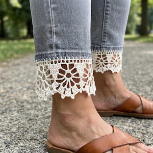 Peut inclure: Jean en denim gris avec une bordure en dentelle crochetée blanc cassé le long de l'ourlet inférieur. Le motif de la dentelle se compose de motifs floraux répétitifs avec des bords festonnés. Le jean est associé à des sandales croisées en cuir marron.