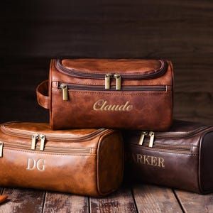 May include: Three brown leather toiletry bags with gold zippers and personalized embroidery. The bags are in varying shades of brown, with the names "Claude", "DG", and "Parker" embroidered on them. The bags are on a wooden surface.