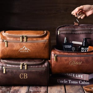 May include: Leather toiletry bags in shades of brown, with gold zippers and personalized embroidery. One bag is open, revealing grooming products. The bags are on a wooden surface, with a book and a small tray.