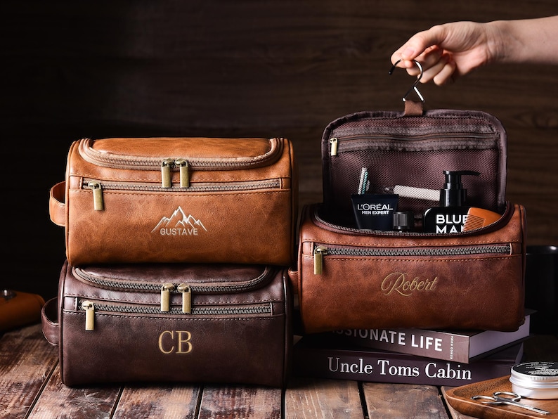 May include: Leather toiletry bags in shades of brown, with gold zippers and personalized embroidery. One bag is open, revealing grooming products. The bags are on a wooden surface, with a book and a small tray.
