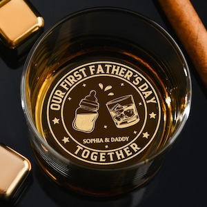 May include: A clear whiskey glass with a gold-colored etched design that reads "OUR FIRST FATHER'S DAY" with a baby bottle and a whiskey glass. The text "SOPHIA & DADDY TOGETHER" is also etched on the glass. Two gold-colored square ice cubes and a cigar are also visible.