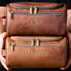 May include: Two brown leather toiletry bags with gold-colored zippers and handles are held up. The top bag has the initials "CNR" embossed on it, while the bottom bag has the name "MARTIN" embossed on it. The bags are a medium brown color.