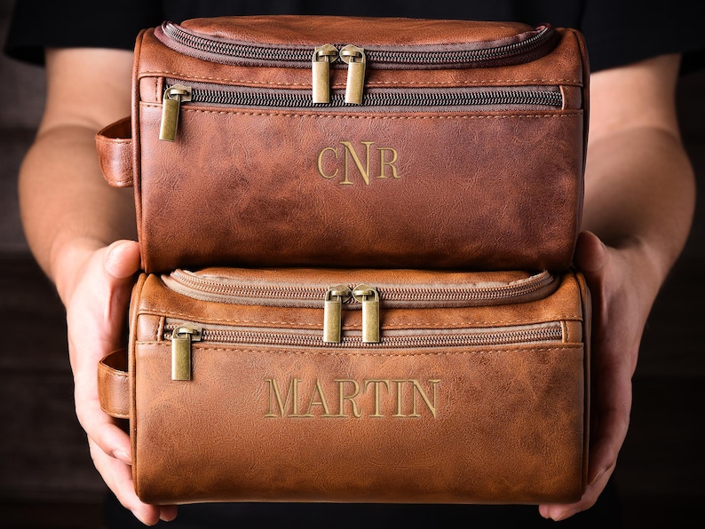 May include: Two brown leather toiletry bags with gold-colored zippers and handles are held up. The top bag has the initials "CNR" embossed on it, while the bottom bag has the name "MARTIN" embossed on it. The bags are a medium brown color.