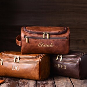May include: Three brown leather toiletry bags with gold zippers and personalized embroidery. The bags are in varying shades of brown, with the names "Claude", "DG", and "Parker" embroidered on them. The bags are on a wooden surface.