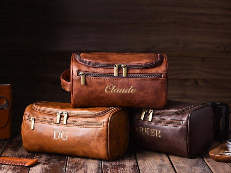 May include: Three brown leather toiletry bags with gold zippers and personalized embroidery. The bags are in varying shades of brown, with the names "Claude", "DG", and "Parker" embroidered on them. The bags are on a wooden surface.
