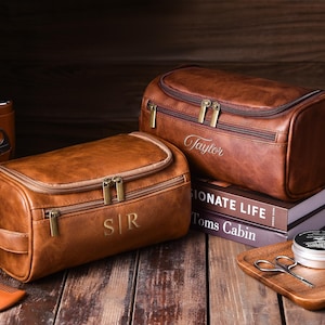 May include: Two brown leather toiletry bags with gold zippers and personalized initials and names. One bag has "S|R" and the other has "Taylor" embroidered on it. The bags are on a wooden surface with grooming accessories.