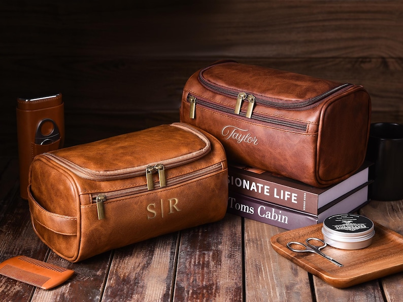 May include: Two brown leather toiletry bags with gold zippers and personalized initials and names. One bag has "S|R" and the other has "Taylor" embroidered on it. The bags are on a wooden surface with grooming accessories.