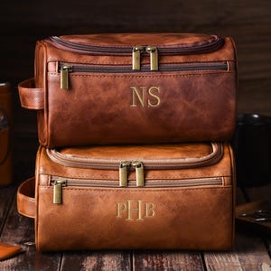 May include: Two stacked brown leather toiletry bags with gold zippers and handles. The top bag has the initials "NS" and the bottom bag has "PHB" embroidered on the front. Other grooming accessories are visible.