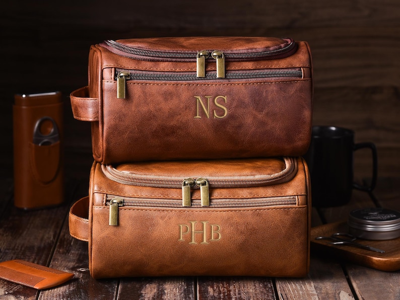 May include: Two stacked brown leather toiletry bags with gold zippers and handles. The top bag has the initials "NS" and the bottom bag has "PHB" embroidered on the front. Other grooming accessories are visible.