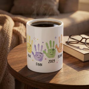 Puede incluir: Taza de cerámica blanca con un diseño de huellas de manos coloridas. La taza presenta huellas de manos en amarillo, morado, verde y naranja, cada una con un nombre debajo. La taza está llena de café oscuro.