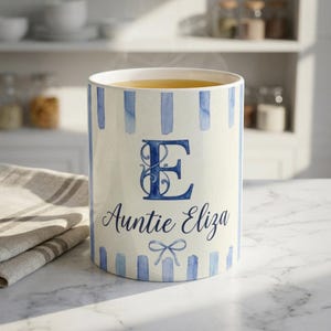 Puede incluir: Una taza de cerámica con fondo color crema y rayas verticales de acuarela azul. El frente muestra una letra E azul grande y ornamentada, el texto Auntie Eliza en letra cursiva y un pequeño gráfico de un lazo azul debajo del nombre.