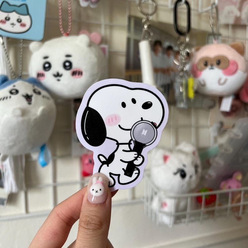 May include: A sticker featuring a cartoon dog with black ears and a pink blush, holding a black microphone with the letters "BTS" on it. The sticker has a white background with a light purple border. Other plush toys and keychains are in the background.