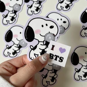 May include: A collection of white stickers featuring a cartoon dog holding a microphone with a purple border. The dog has black ears and a pink blush. A small square sticker with "I BTS" and a purple heart is held in the hand.