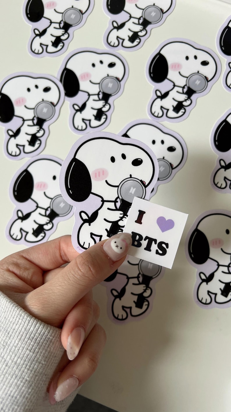 May include: A collection of white stickers featuring a cartoon dog holding a microphone with a purple border. The dog has black ears and a pink blush. A small square sticker with "I BTS" and a purple heart is held in the hand.
