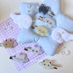 May include: A collection of whimsical hair clips featuring various animal designs with angel wings. The clips are made of clear acrylic and have gold-colored metal clasps. They are arranged on a light blue star-shaped pillow and a checkered pink and white surface.