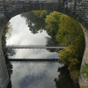 Pu&ograve; includere: Un ponte ad arco in pietra incornicia la vista di un fiume calmo che riflette il cielo e gli alberi. Un ponte sospeso in metallo attraversa l'acqua, creando una doppia immagine nel riflesso. Fogliame verde e giallo costeggia le rive del fiume.