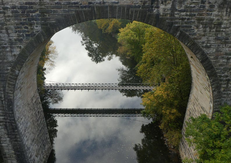 Pu&ograve; includere: Un ponte ad arco in pietra incornicia la vista di un fiume calmo che riflette il cielo e gli alberi. Un ponte sospeso in metallo attraversa l'acqua, creando una doppia immagine nel riflesso. Fogliame verde e giallo costeggia le rive del fiume.