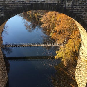 Pu&ograve; includere: Un ponte ad arco in pietra incornicia la vista di un fiume calmo che riflette il cielo azzurro. Una stretta passerella pedonale in metallo attraversa l'acqua, circondata da alberi con fogliame autunnale nei toni del giallo, arancio e marrone.