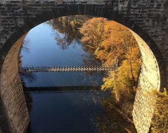 May include: A stone arch bridge frames a view of a calm river reflecting the blue sky. A narrow metal pedestrian bridge spans the water, surrounded by trees with autumn foliage in shades of yellow, orange, and brown under the stone structure.