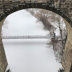 Pu&ograve; includere: Un ponte ad arco in pietra incornicia la vista di un paesaggio innevato con un lungo e sottile ponte sospeso che attraversa un fiume bianco e ghiacciato, fiancheggiato da alberi spogli durante una nevicata.