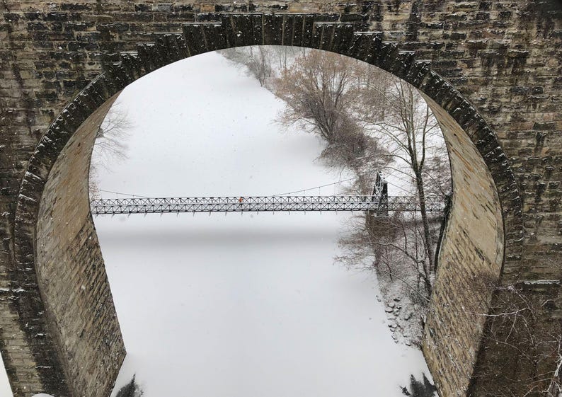 Pu&ograve; includere: Un ponte ad arco in pietra incornicia la vista di un paesaggio innevato con un lungo e sottile ponte sospeso che attraversa un fiume bianco e ghiacciato, fiancheggiato da alberi spogli durante una nevicata.