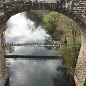 Pu&ograve; includere: Un ponte ad arco in pietra incornicia la vista di un fiume calmo sottostante. Una stretta passerella sospesa in metallo attraversa l'acqua orizzontalmente. Il paesaggio circostante presenta alberi verdi e riflessi del cielo sull'acqua.