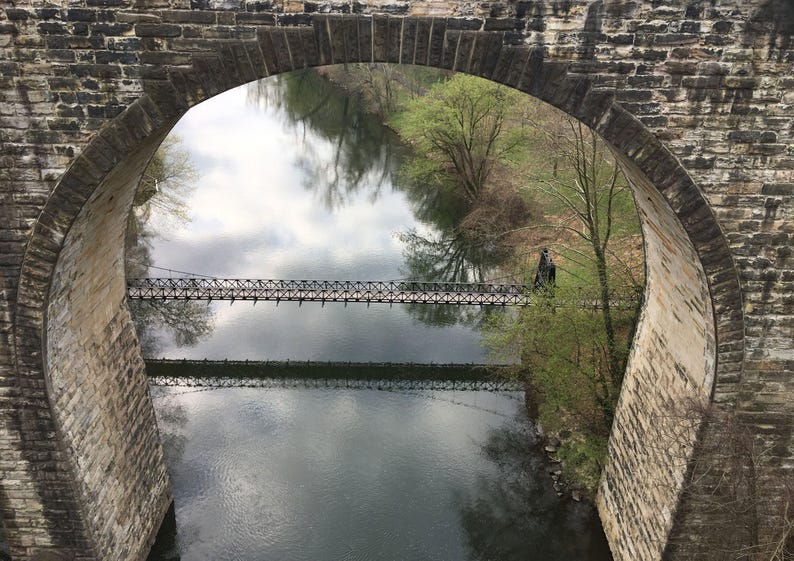 Pu&ograve; includere: Un ponte ad arco in pietra incornicia la vista di un fiume calmo sottostante. Una stretta passerella sospesa in metallo attraversa l'acqua orizzontalmente. Il paesaggio circostante presenta alberi verdi e riflessi del cielo sull'acqua.