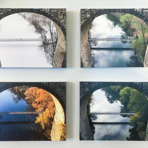 Pu&ograve; includere: Quattro stampe rettangolari su tela disposte a griglia. Ogni stampa presenta un arco in pietra che incornicia la vista di un ponte sospeso su un fiume, mostrando lo stesso paesaggio durante le quattro diverse stagioni dell'anno.