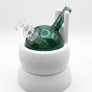 May include: A dark green glass water pipe with a clear glass bowl piece, resting inside a matte white, two-tiered cylindrical stand. The stand features two vertical prongs that extend upward to support the neck of the glass pipe securely.