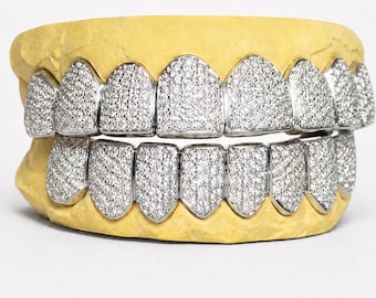 Op de afbeelding: Een set zilverkleurige grillz met een dichte pavé-zetting van kleine, heldere, reflecterende steentjes die het volledige oppervlak van elke tand bedekken. Het gebitsaccessoire is bevestigd op een gele, getextureerde gebitsafdruk.