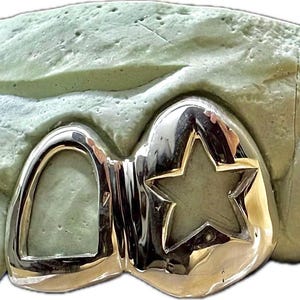 May include: Silver-colored dental grill with a star-shaped cutout, displayed on a light green dental mold. The grill has a polished, reflective surface, showcasing a custom design for tooth enhancement.
