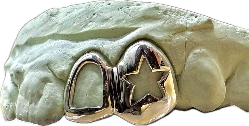 May include: Silver-colored dental grill with a star-shaped cutout, displayed on a light green dental mold. The grill has a polished, reflective surface, showcasing a custom design for tooth enhancement.