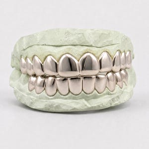 Puede incluir: Un conjunto de fundas dentales de metal pulido en color oro rosa, ajustadas sobre un molde dental de color verde claro. Las fundas cubren las filas superior e inferior de dientes, con una superficie lisa y reflectante que imita la forma natural.