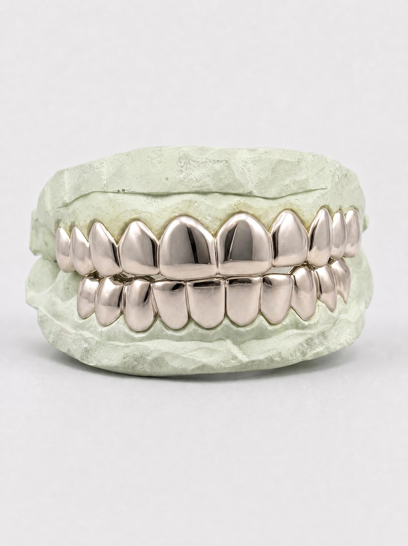 Puede incluir: Un conjunto de fundas dentales de metal pulido en color oro rosa, ajustadas sobre un molde dental de color verde claro. Las fundas cubren las filas superior e inferior de dientes, con una superficie lisa y reflectante que imita la forma natural.