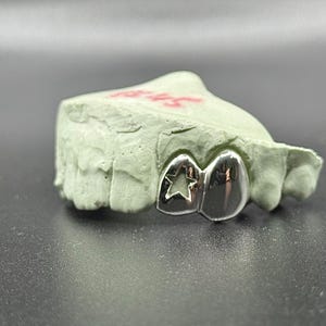 May include: A silver dental grill with a star cutout design, resting on a light green dental mold. The grill has a polished, reflective surface. The mold has red markings.