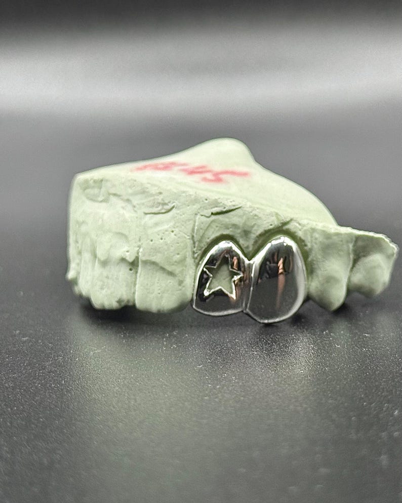 May include: A silver dental grill with a star cutout design, resting on a light green dental mold. The grill has a polished, reflective surface. The mold has red markings.