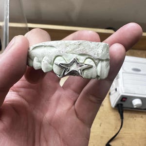 May include: A silver star-shaped grill is set on a dental mold. The grill is a five-pointed star with a polished finish. The mold is a light green color and has several tooth impressions.