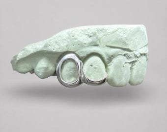 Puede incluir: Una pieza de grillz de plata pulida ajustada sobre dos dientes en un molde dental de color verde claro. El accesorio de metal presenta una superficie lisa y reflectante que se adapta con precisión a la forma de los dientes, mostrando un acabado metálico.