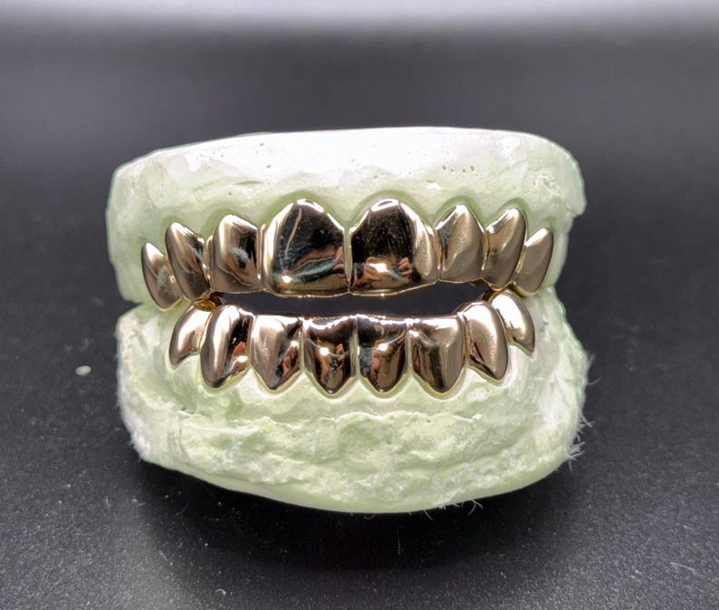 Puede incluir: Un juego de fundas dentales de metal en tono dorado pulido ajustadas sobre un molde dental de color verde claro. Las fundas cubren las filas superior e inferior de los dientes, con una superficie lisa y reflectante que imita la forma natural.