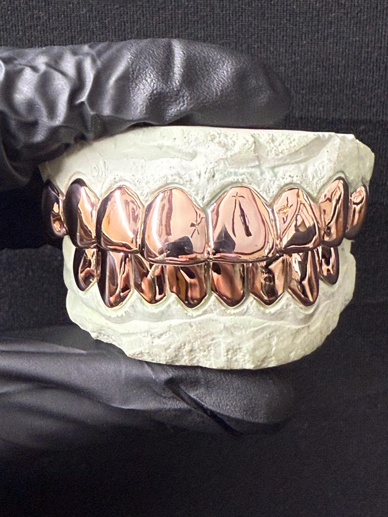 Puede incluir: Un conjunto de fundas dentales de metal color oro rosa ajustadas sobre un molde dental blanco. La superficie met&aacute;lica es altamente reflectante y cubre tanto la fila superior como la inferior de los dientes, con un acabado liso y pulido.