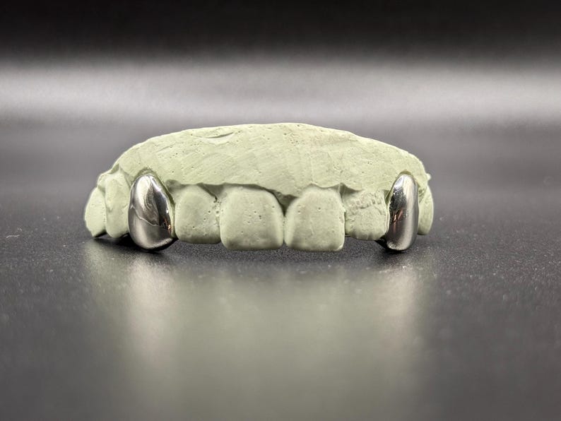 Puede incluir: Tapones dentales plateados expuestos sobre un molde dental verde claro. Los tapones tienen una superficie pulida y reflectante, que contrasta con la textura mate del molde. Los tapones est&aacute;n dise&ntilde;ados para adaptarse a los dientes delanteros.