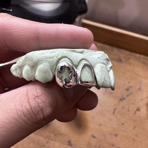 May include: Silver dental grill with a star-shaped design on one tooth and a smooth surface on the other. The grill is set on a green dental mold, held by a hand. The background is a wooden surface.