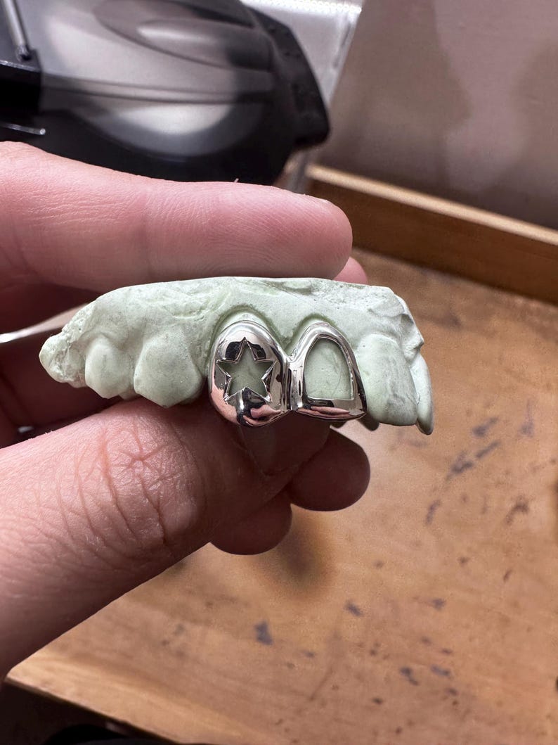 May include: Silver dental grill with a star-shaped design on one tooth and a smooth surface on the other. The grill is set on a green dental mold, held by a hand. The background is a wooden surface.