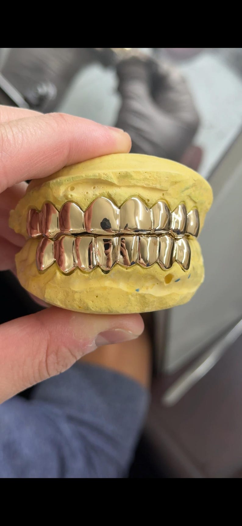 Puede incluir: Un conjunto de parrillas dentales de color oro pulido ajustadas sobre los dientes superiores e inferiores de un molde dental amarillo. La parrilla met&aacute;lica cubre ocho dientes en la fila superior y ocho en la inferior, reflejando luz en su superficie.