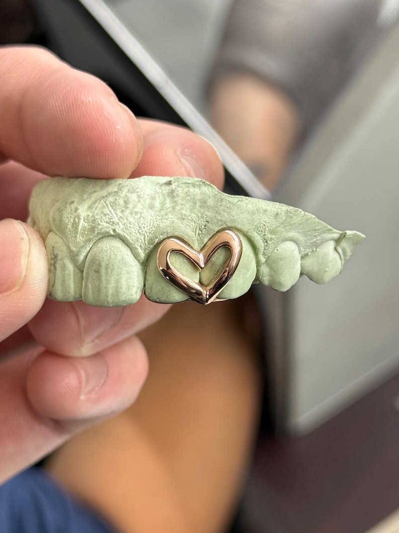 May include: A gold-colored heart-shaped tooth grill is set on a green dental mold. The heart grill is a delicate, open design, showcasing a polished finish. The mold provides a clear view of the grill's fit and form.