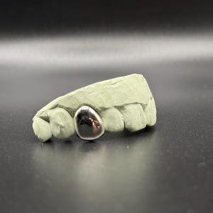 Puede incluir: Una impresi&oacute;n dental con una sola corona dental plateada pulida. La corona es lisa y reflectante, contrastando con el molde dental texturizado, verde claro. El fondo es de un tono oscuro y neutro.