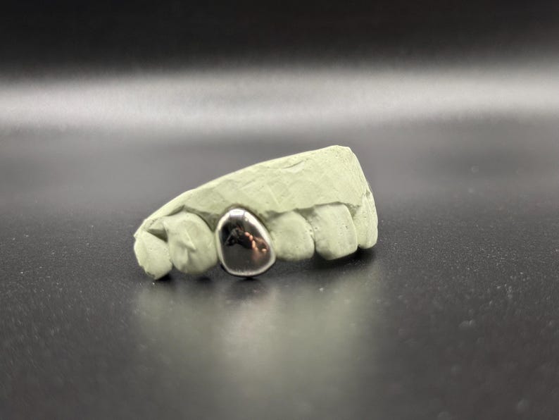 Puede incluir: Una impresi&oacute;n dental con una sola corona dental plateada pulida. La corona es lisa y reflectante, contrastando con el molde dental texturizado, verde claro. El fondo es de un tono oscuro y neutro.